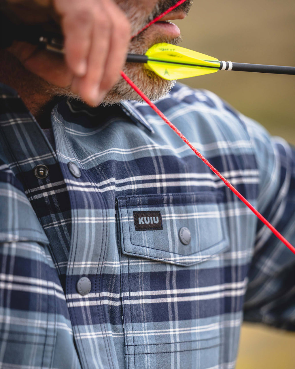 Close-up of a man aiming a bow with an arrow nocked against his lips. He is wearing KUIU Field Flannel Fleece in Steel Blue Framework showing a chest pocket featuring a black KUIU label.