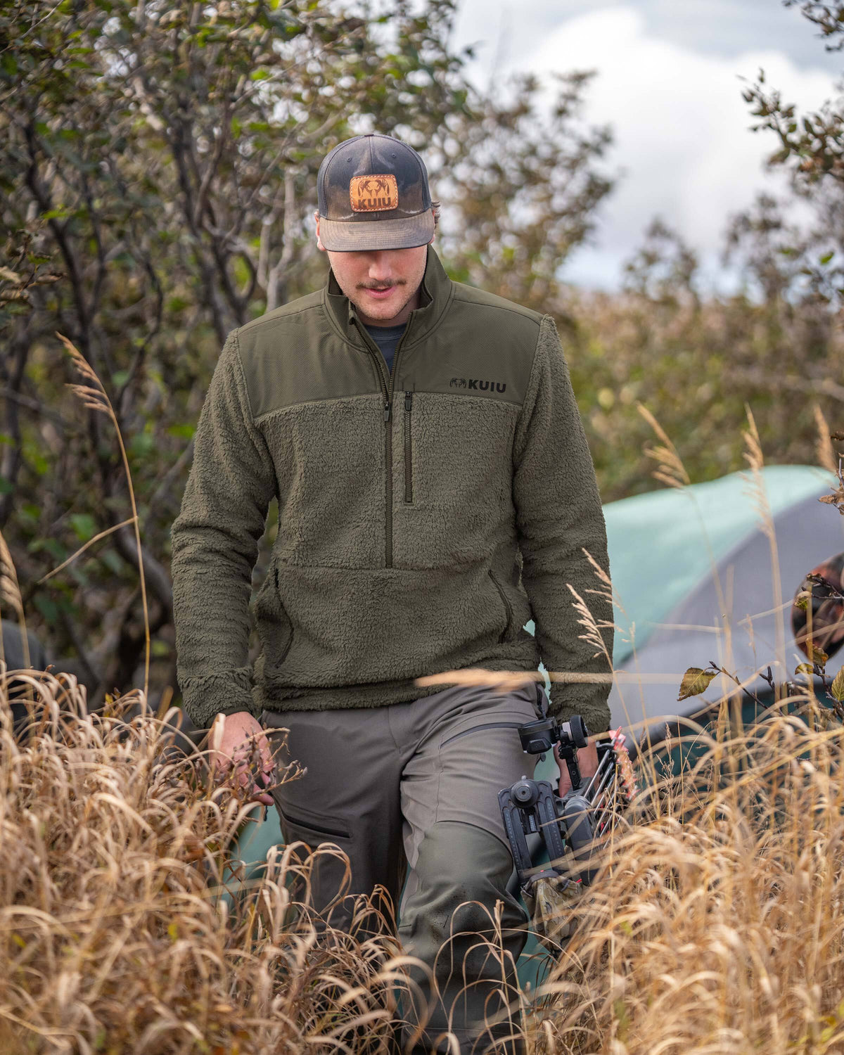 Hunter wearing Sherpa Fleece Zip Pullover in Ash Brown, holding a compound bow, walking through tall dried grass 