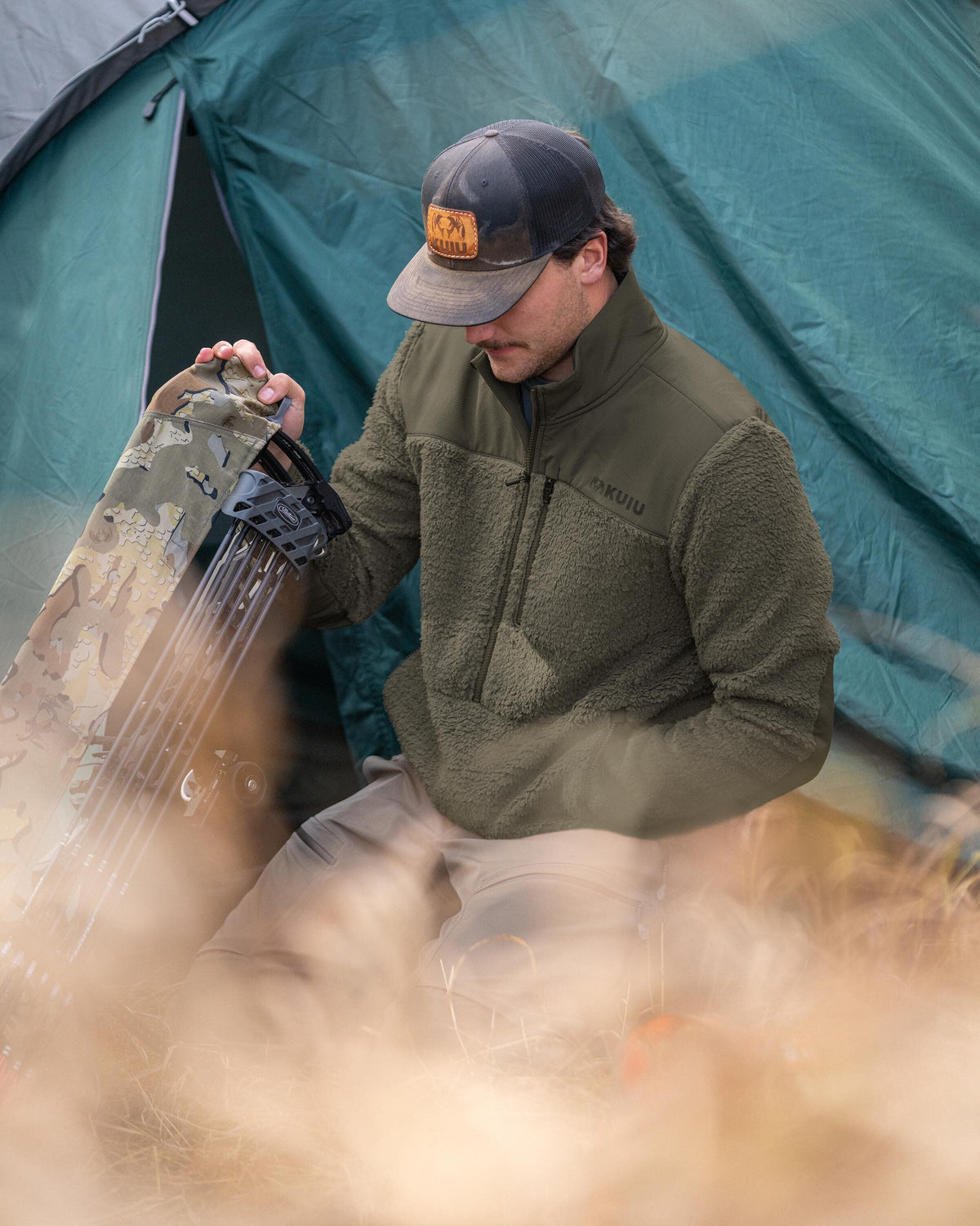 Hunter wearing Sherpa Fleece Zip Pullover in Ash Brown sitting in front of a blueish green tent next to a compound bow