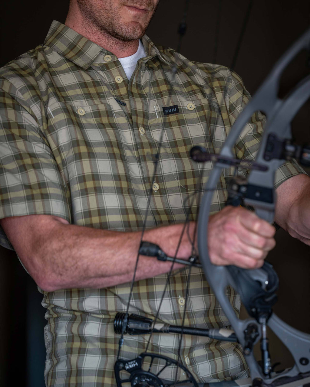 Close-up of a man wearing Borrego Short Sleeve Shirt in Valo Crosscheck Plaid adjusting the grip and cables of a compound bow.