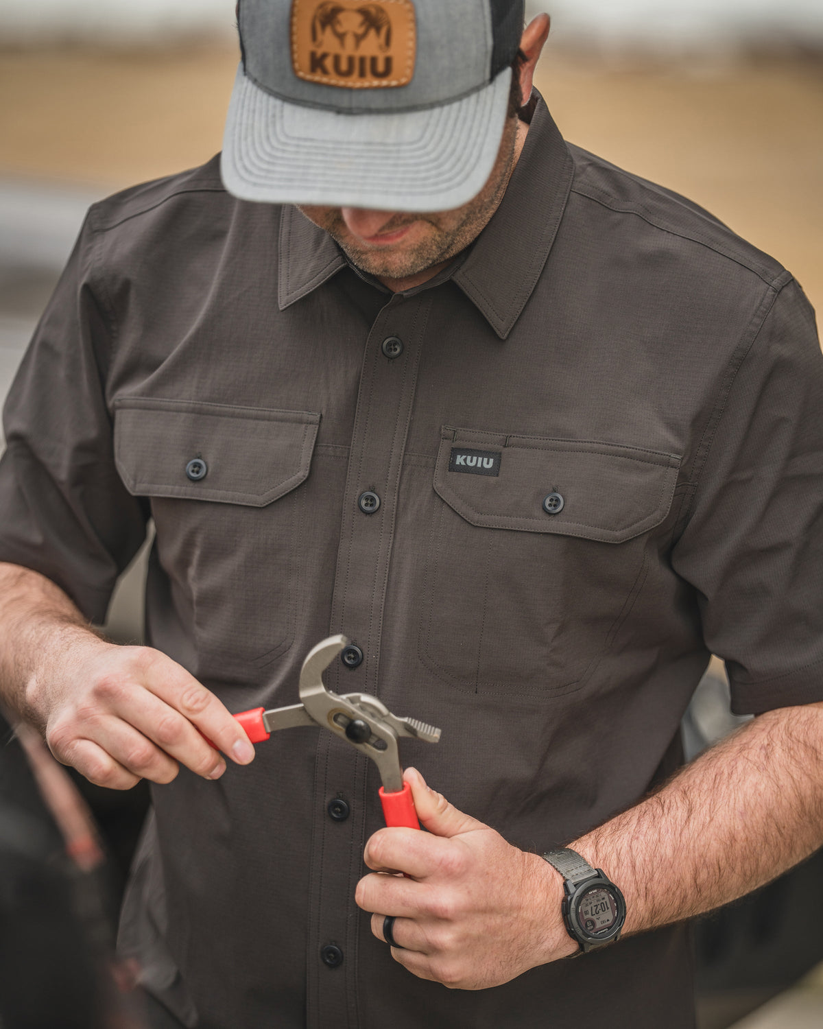 Man wearing Gauge Short Sleeve Work Shirt in Rodeo Brown holding red‑handled pliers while working near a vehicle outdoors.