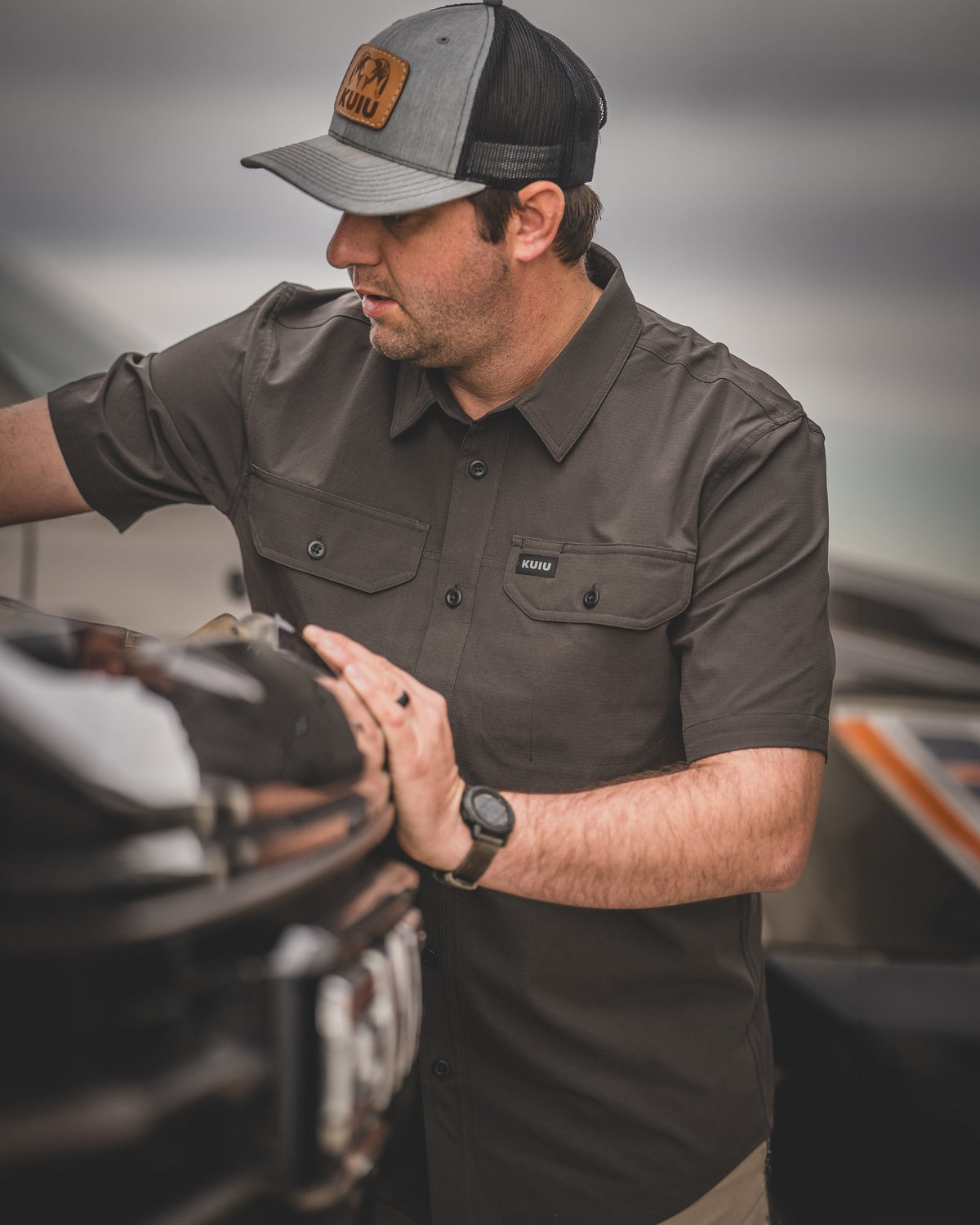 Man wearing Gauge Short Sleeve Work Shirt in Rodeo Brown leaning over the front of a vehicle with hands positioned on the hood.