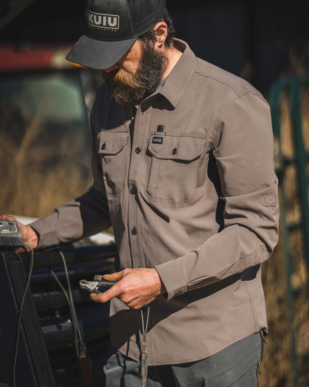 Man wearing a Gauge Long Sleeve Work Shirt in Rodeo Brown standing beside a vehicle and holding a metal clip and cord.