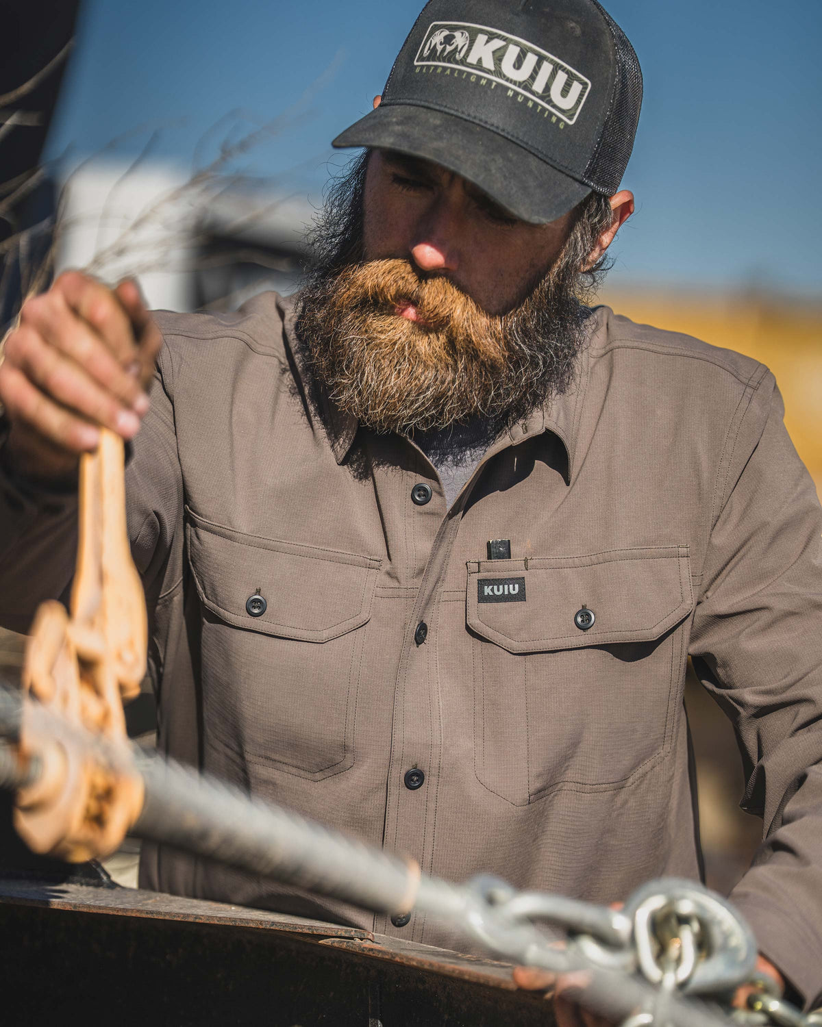 Man wearing Gauge Long Sleeve Work Shirt in Rodeo Brown tightening a metal hardware outdoors.