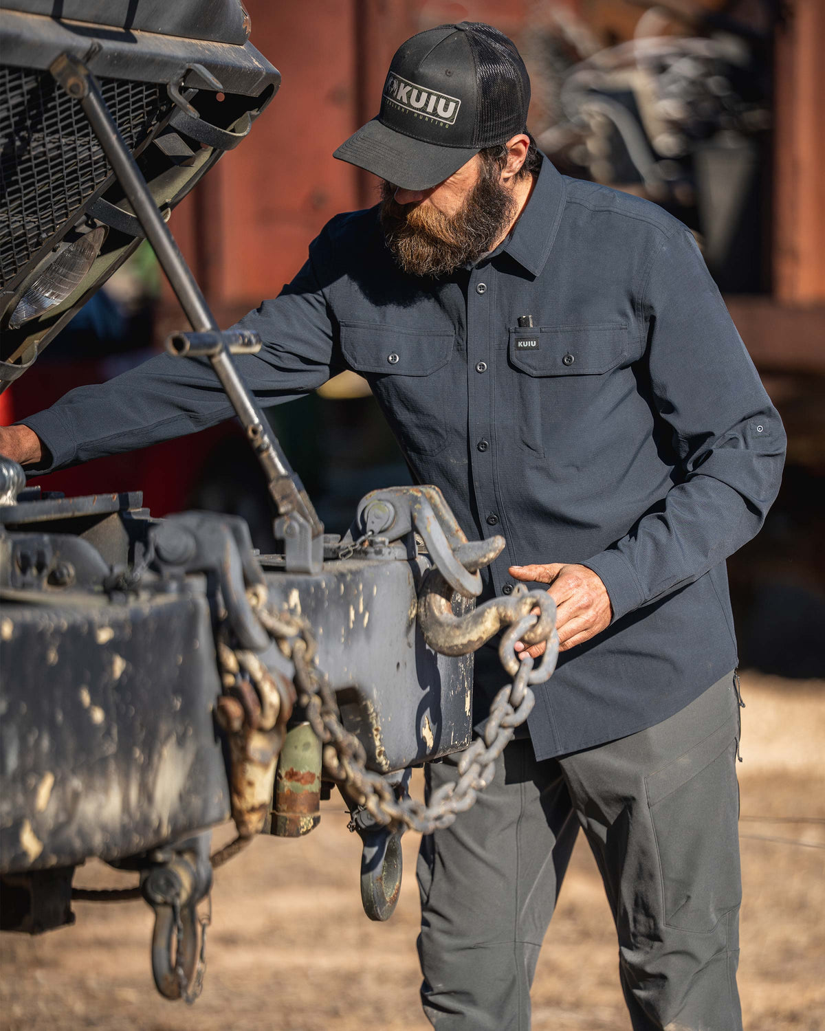 Man wearing Gauge Long Sleeve Work Shirt in Nightfall Blue, working on an open vehicle engine and handling a chain outdoors near rust‑colored freight cars.