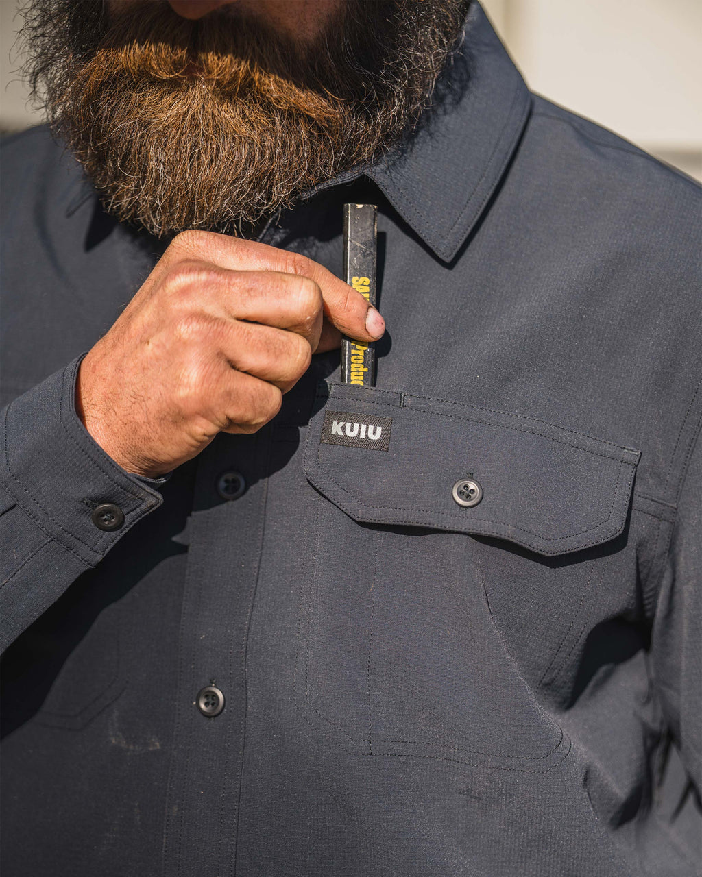 Close‑up of a person placing a rectangular tool or marker into the pencil divider pocket of the Gauge Long Sleeve Work Shirt in Nighfall Blue.