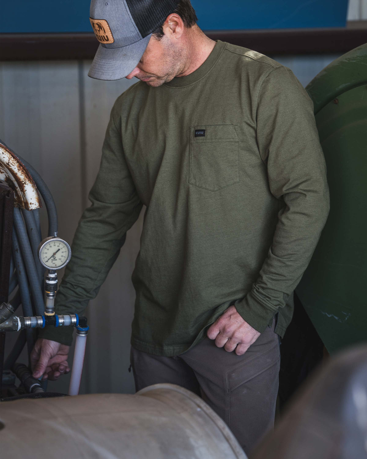 Man in an industrial workspace, wearing Workgrade Long Sleeve Pocket Crew in Evergreen, standing next to a set of hoses connected to pressure gauge.