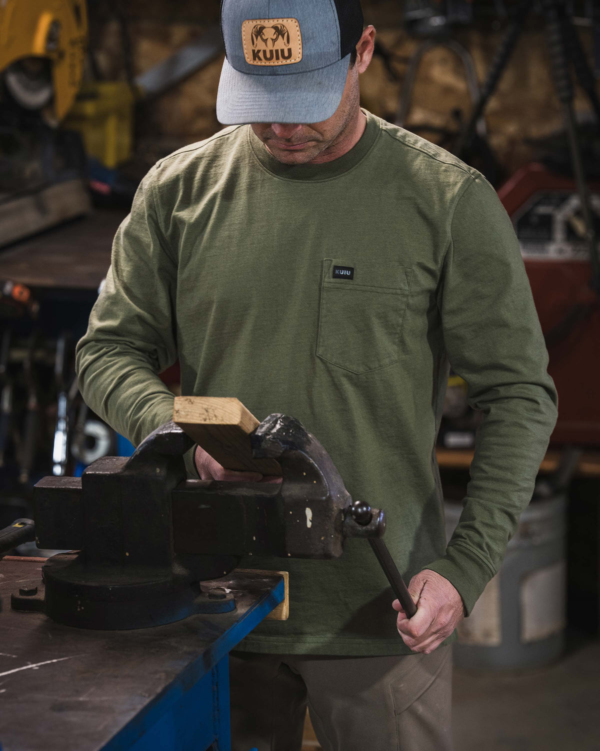 Man wearing Workgrade Long Sleeve Pocket Crew, standing behind a workbench, working with a heavy metal bench vise mounted to the work surface.