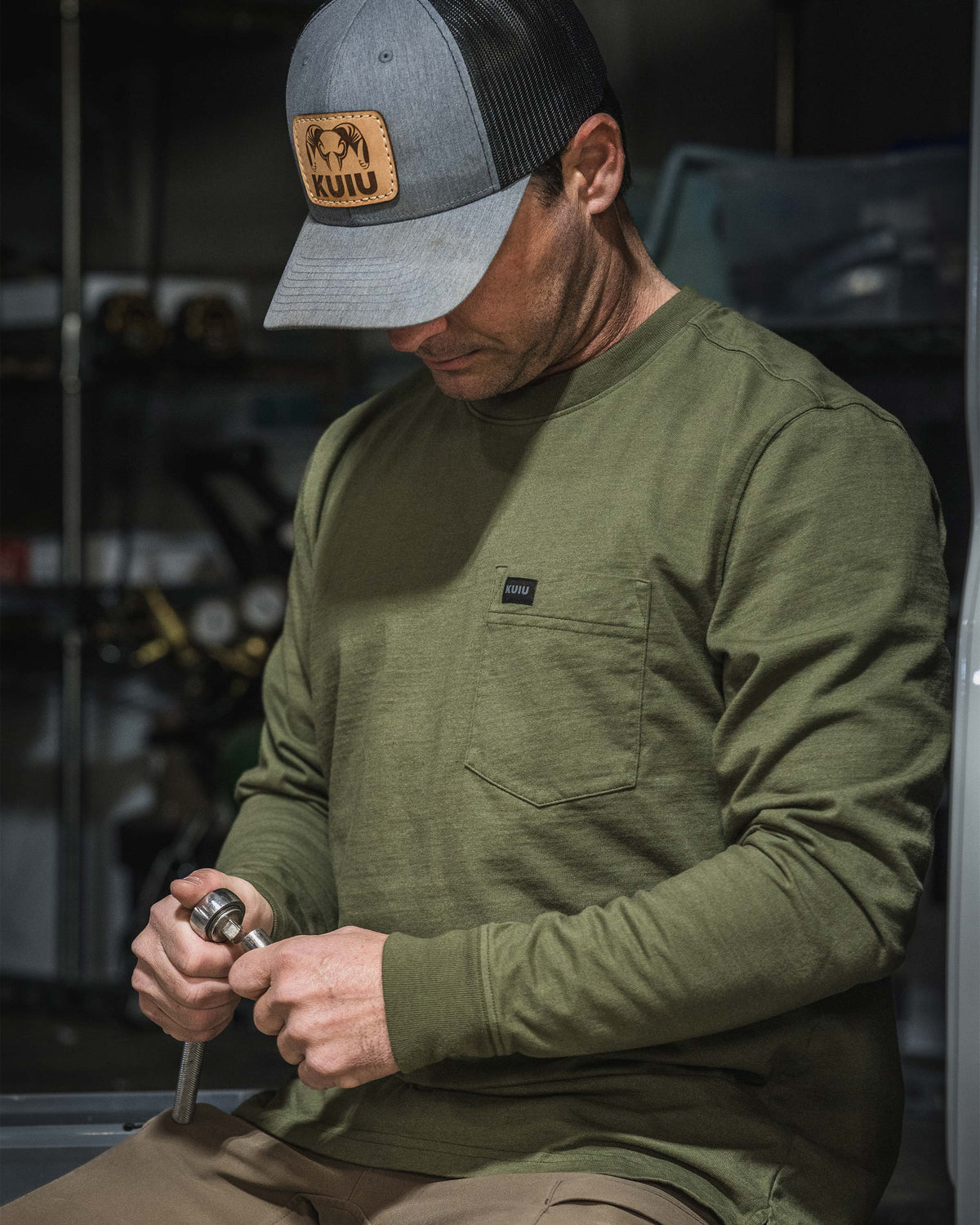 Man sitting in a workshop, holding a socket wrench tool, wearing Workgrade Long Sleeve Pocket Crew in Evergreen.