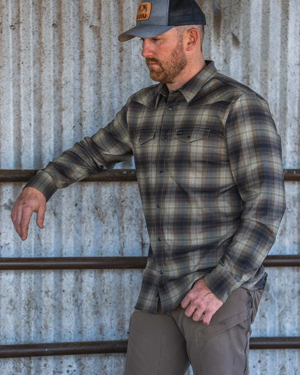Man standing against a corrugated metal wall, wearing a Laredo Long Sleeve Shirt in Vias Ombre Plaid in an outdoor ranch setting.

