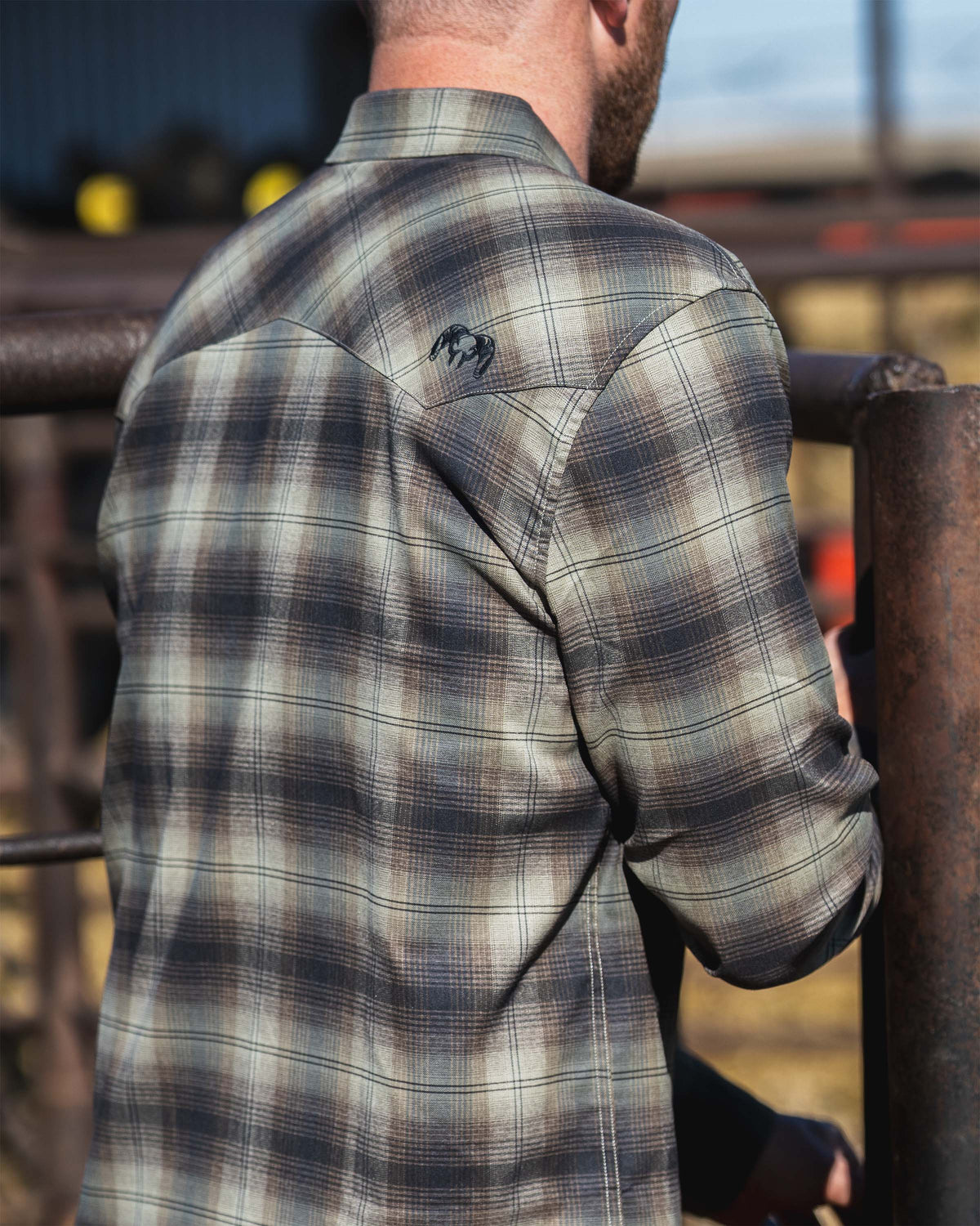 Back view of a man wearing a Laredo Long Sleeve Shirt in Vias Ombre Plaid, showing the shoulder yoke and embroidered KUIU logo detail while standing near a metal fence.
