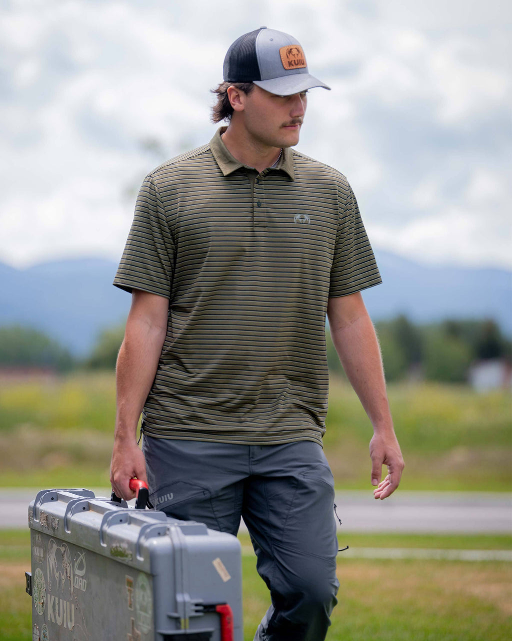 Man wearing a KUIU Performance Polo in Evergreen Stripe walking in an outdoor setting while carrying a gray hard case.