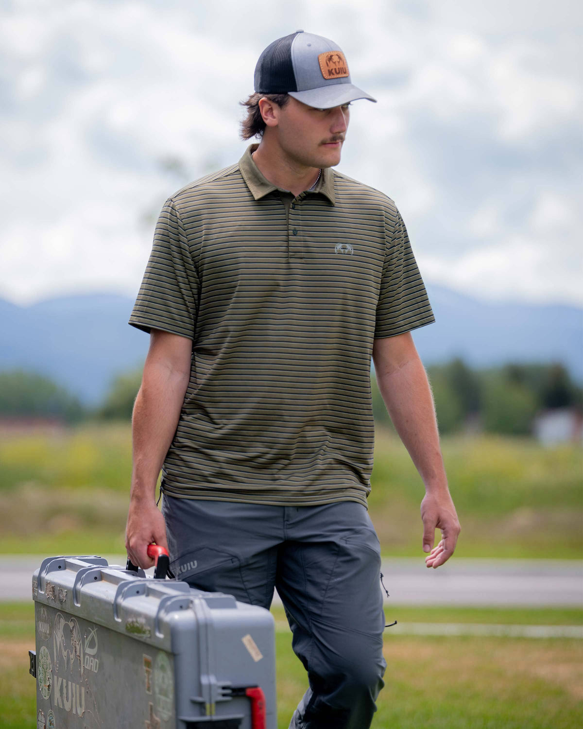 Man wearing a KUIU Performance Polo in Evergreen Stripe walking in an outdoor setting while carrying a gray hard case.