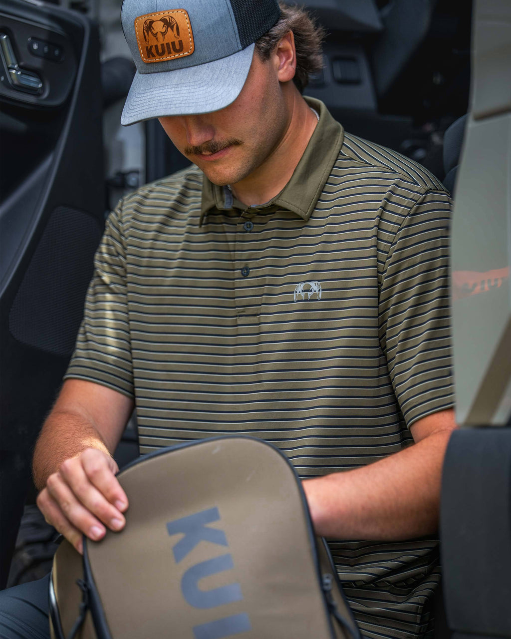 Man wearing a KUIU Performance Polo in Evergreen Stripe sits inside a vehicle rummaging through a KUIU Waypoint Travel Backpack in Bourbon Brown.