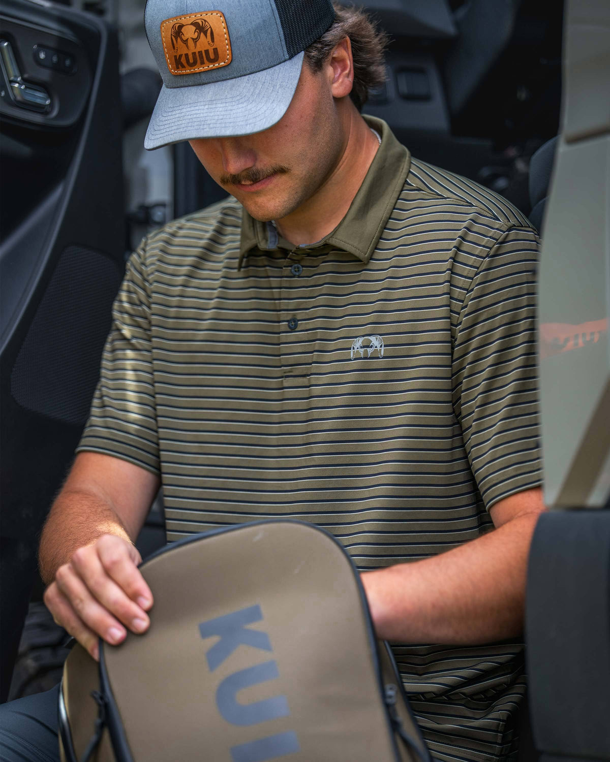 Man wearing a KUIU Performance Polo in Evergreen Stripe sits inside a vehicle rummaging through a KUIU Waypoint Travel Backpack in Bourbon Brown.