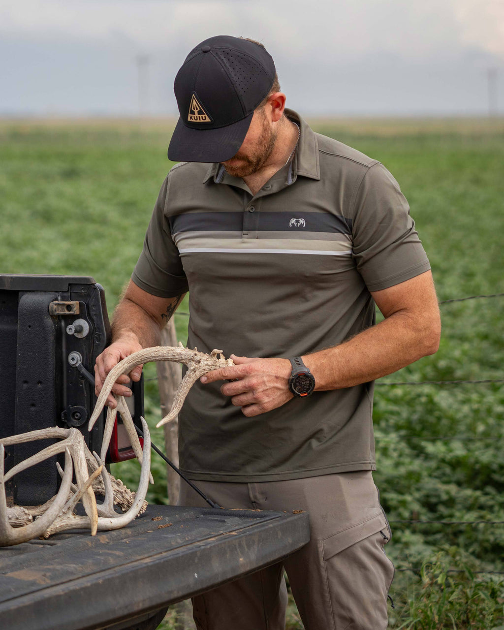 Man in an outdoor setting standing by a pickup truck examining deer antlers wearing Performance Polo in Ash Chest Stripe