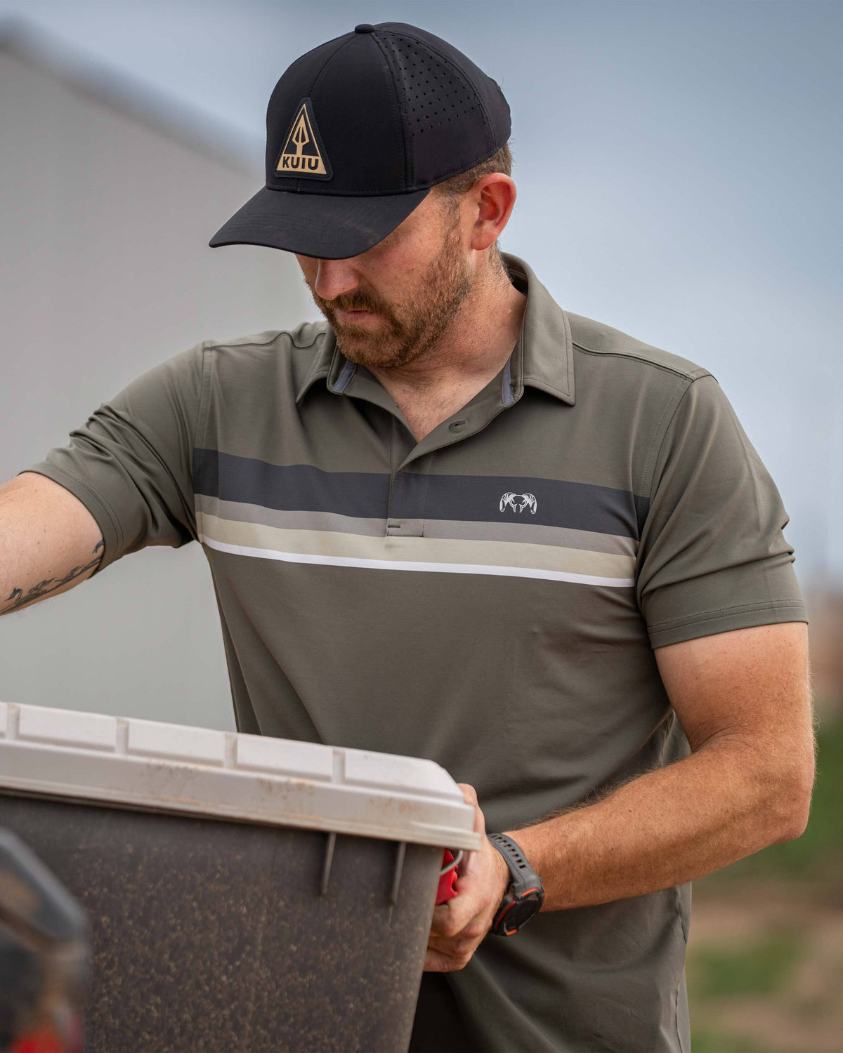 Man lifting a storage chest from a pickup truck wearing KUIU Performance Polo Chest Stripe in Ash Stripe