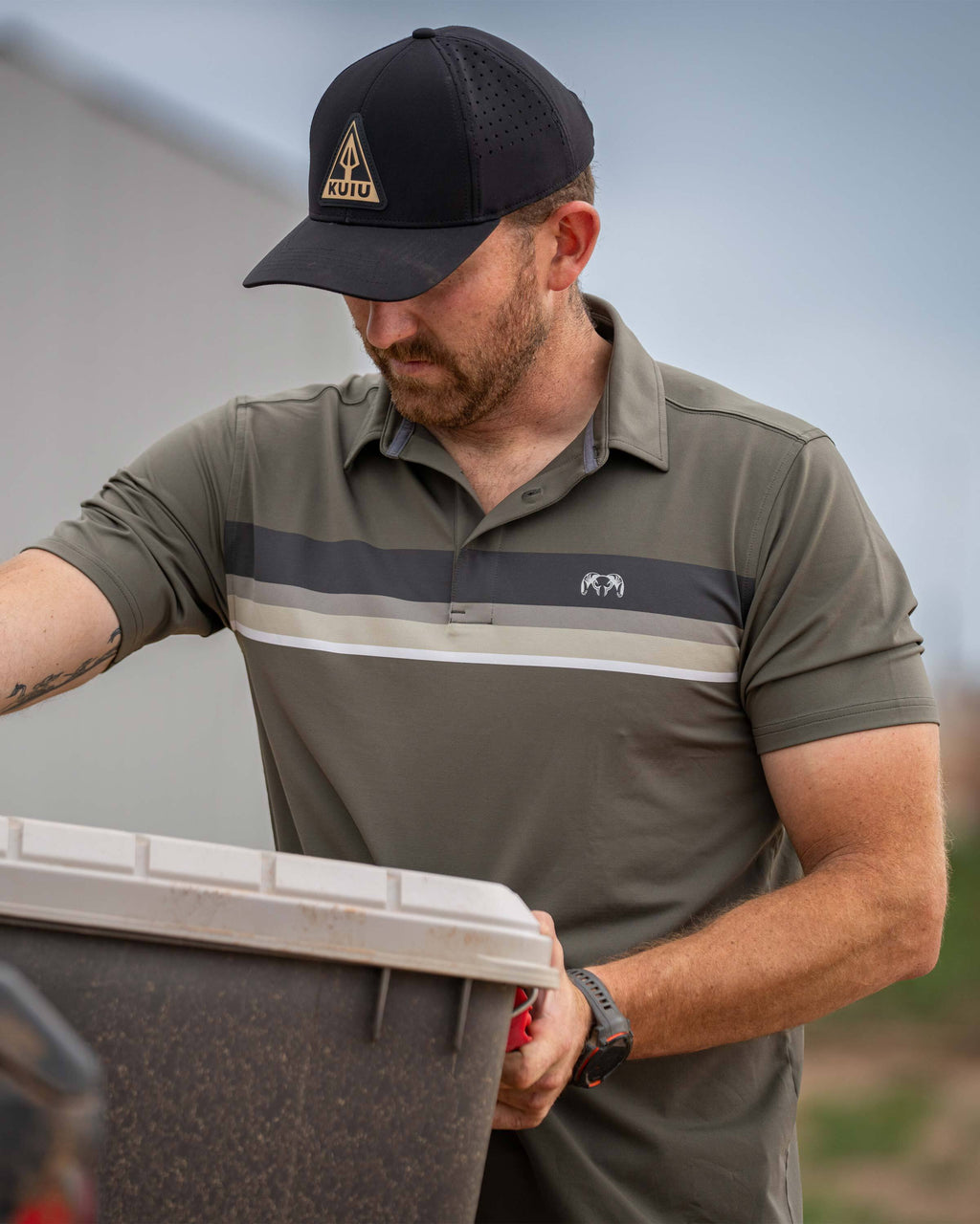Man lifting a storage chest from a pickup truck wearing KUIU Performance Polo Chest Stripe in Ash Stripe