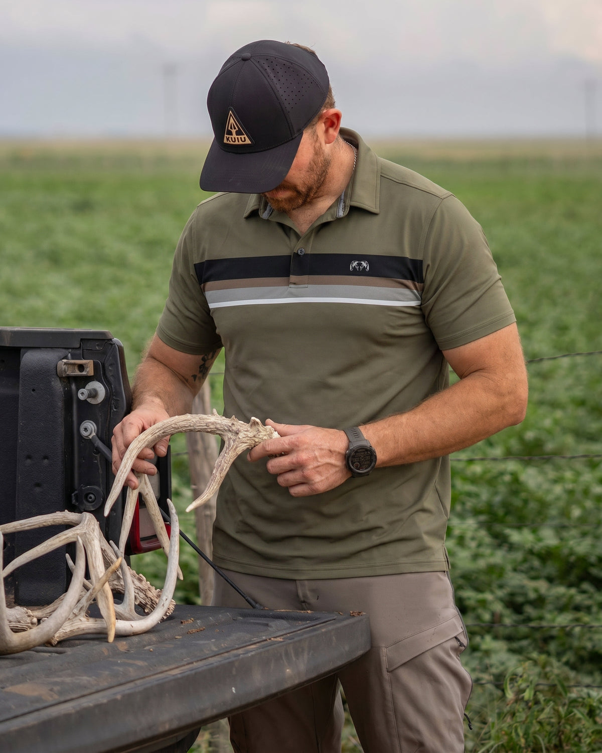 Man in an outdoor setting standing by a pickup truck examining deer antlers wearing Performance Polo in Evergreen Chest Stripe