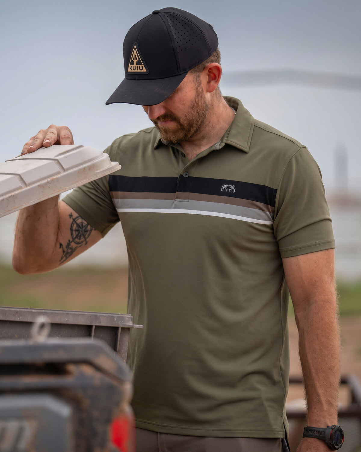 Man lifts storage chest lid and appears to look inside wearing KUIU Performance Polo Chest Stripe in Evergreen Chest Stripe