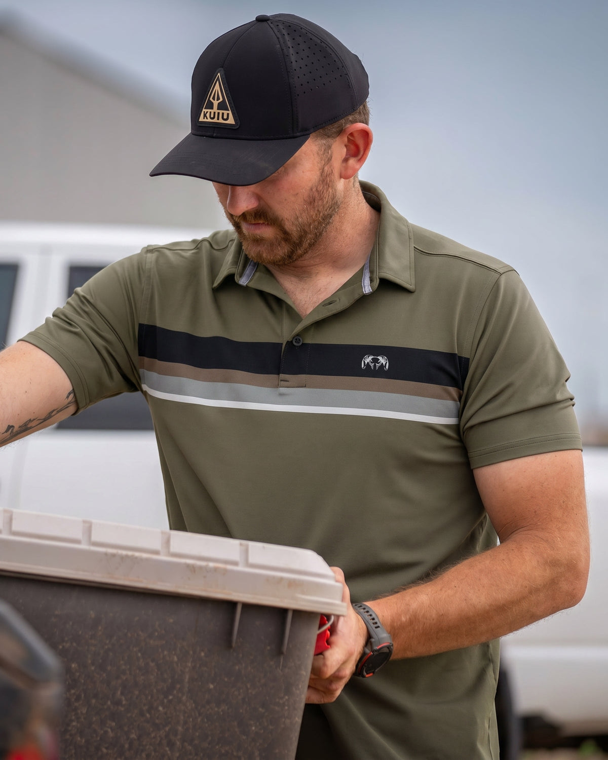 Man lifts storage chest lid and appears to look inside wearing KUIU Performance Polo Chest Stripe in Evergreen Chest Stripe