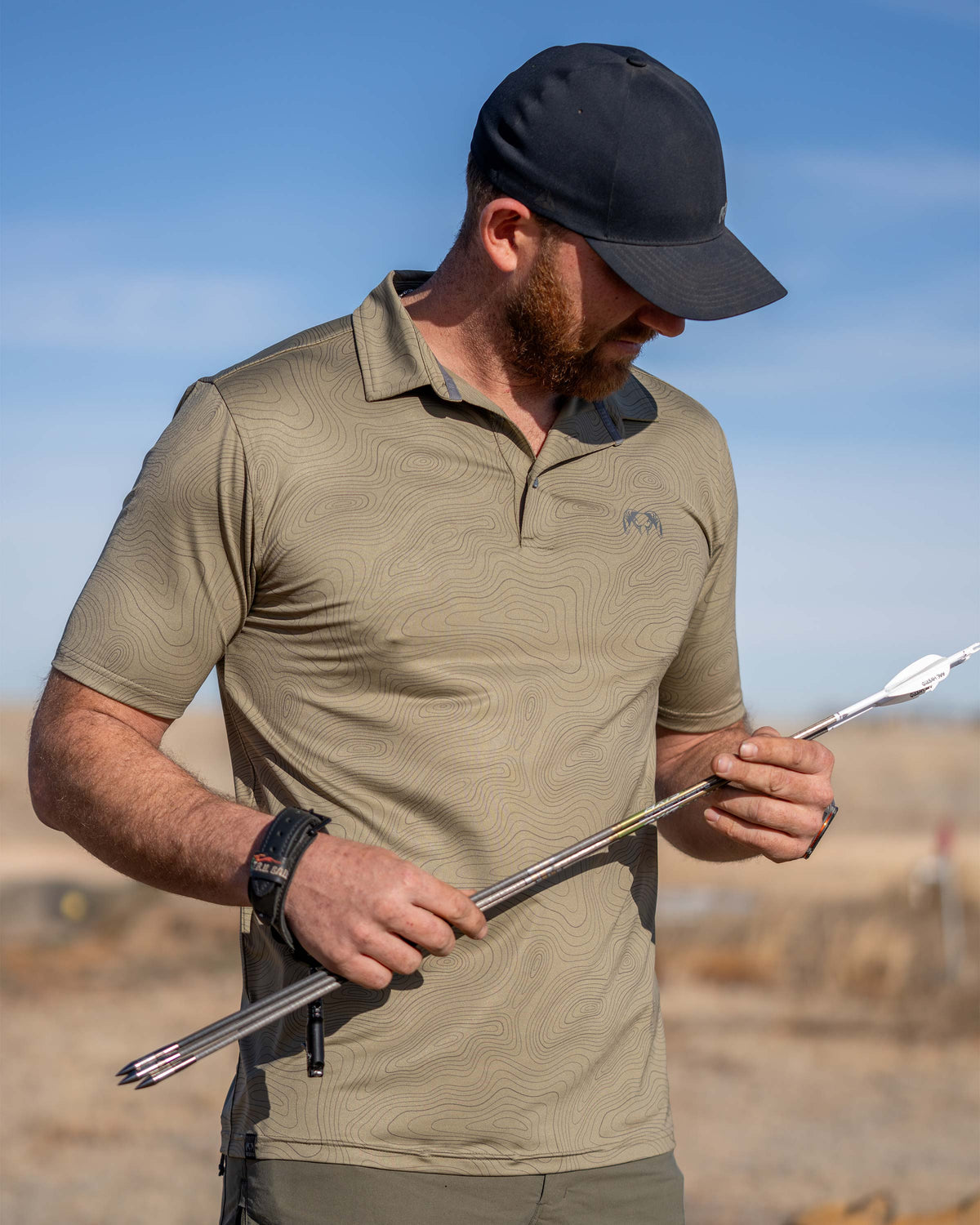 Hunter holding and looking down at his arrows, wearing KUIU Performance Polo in Khaki Topo