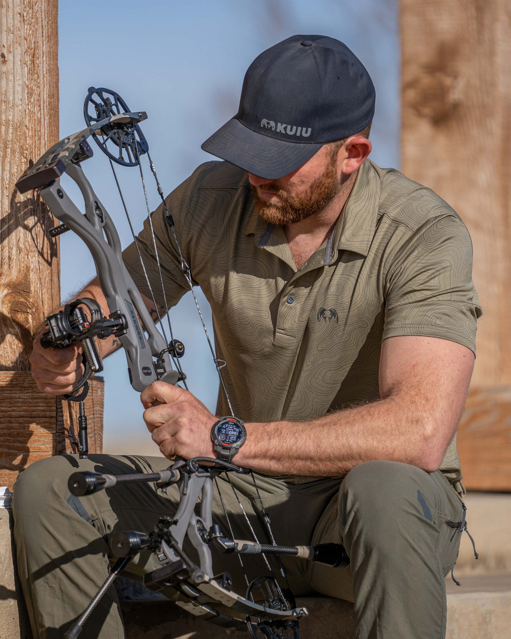 Hunter sitting and adjusting his bow, wearing KUIU Performance Polo in Khaki Topo