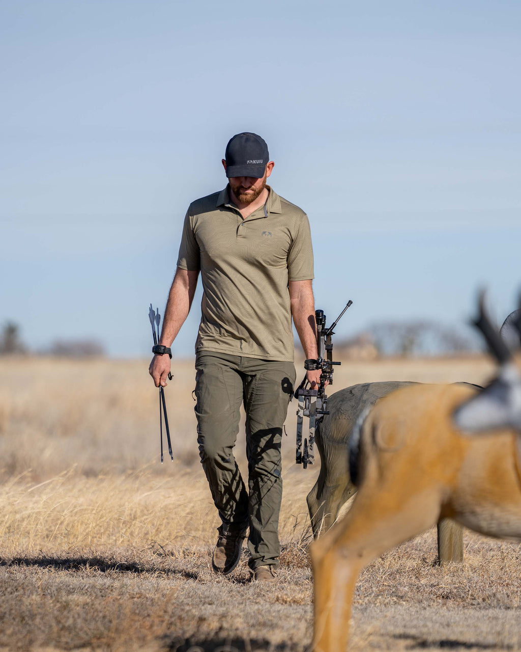 Hunter walking through a field with a couple deer decoys, carrying his bow and arrows, wearing KUIU Performance Polo in Khaki Topo