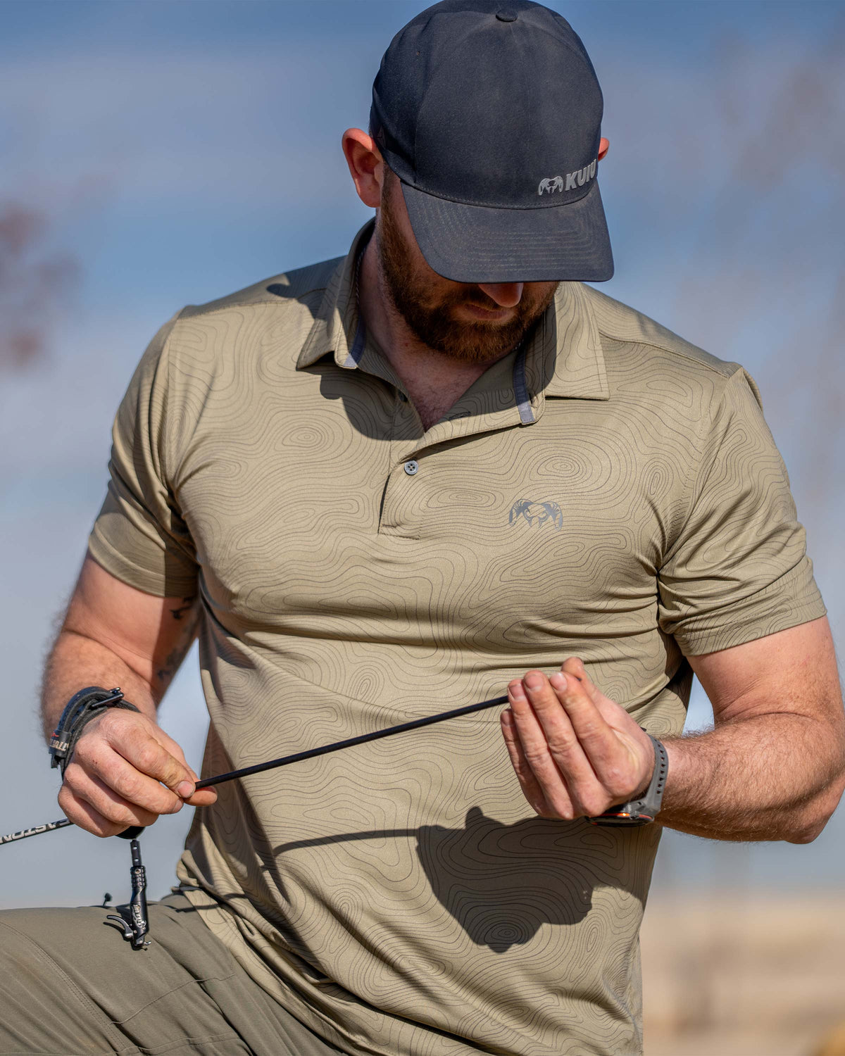 Hunter viewing the tip of his arrow, wearing KUIU Performance Polo in Khaki Topo