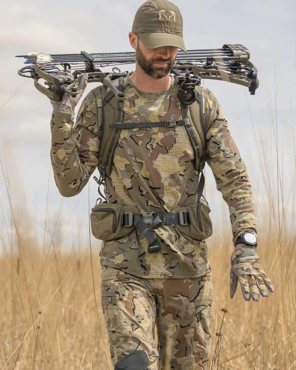 Hunter walking throught tall dry grass with bow over shoulder wearing ULTRA Merino 120 LT LS Crew-T in Valo Camouflage