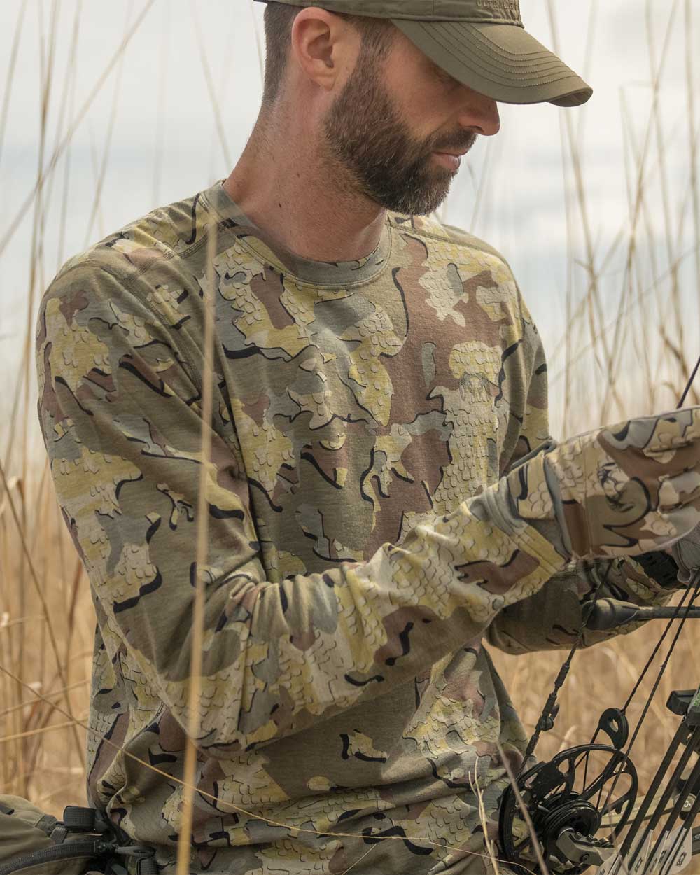 Hunter kneeling in tall dry grass while adjusting bow wearing ULTRA Merino 120 LT LS Crew-T in Valo Camouflage