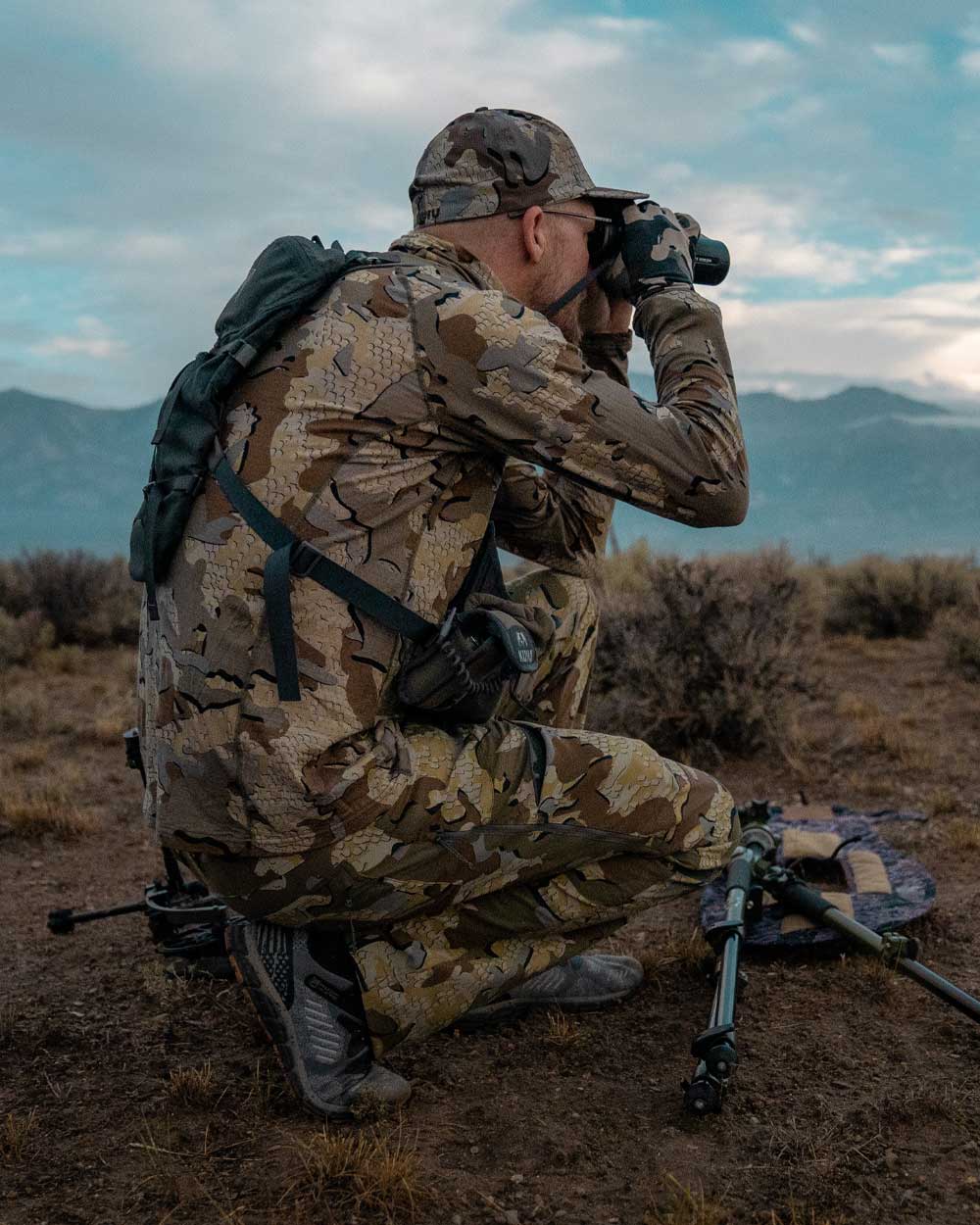 Hunter kneeling in open sage brush terrain, glassing for antelope while wearing ULTRA Merino 120 LT LS Crew-T in Valo Camouflage, side facing view