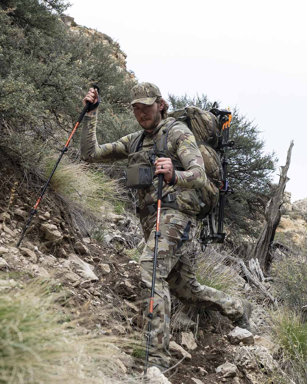 Hunter traversing hill wearing ULTRA Merino 120 LT LS Hoodie in Valo Camouflage