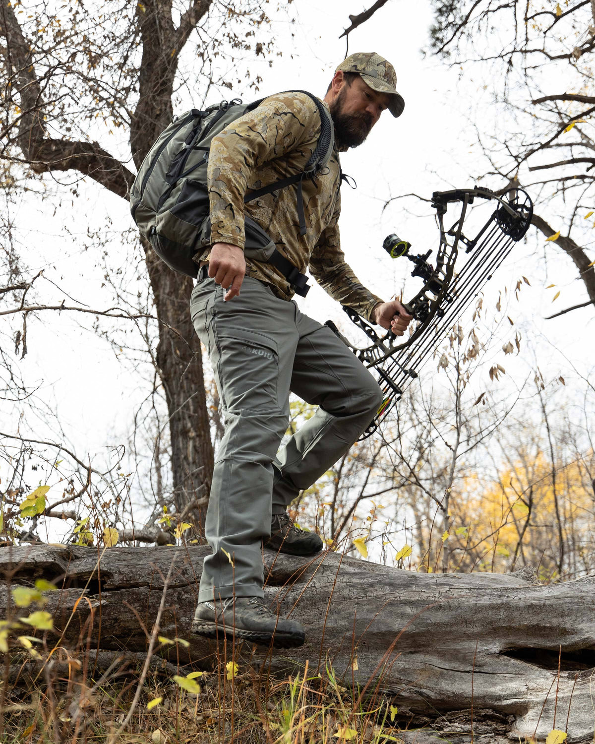 Hunter backpacking, stepping off a wooden log, wearing Ultra Merino 145 Zip-T Hunting Hoodie in Valo Camouflage