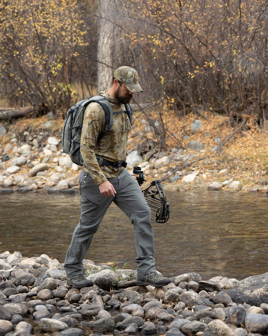 Hunter hiking by a stream, holding a bow, wearing ULTRA Merino 145 Zip-T Hunting Hoodie in Valo Camouflage