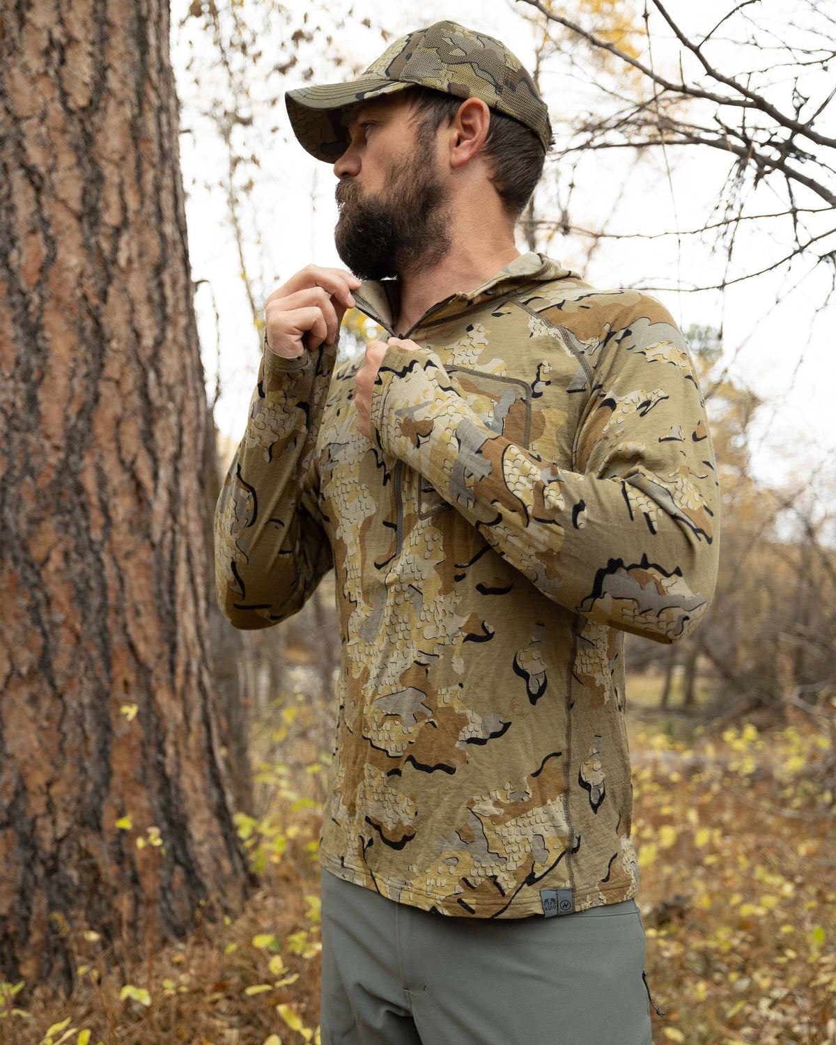 Hunter looking away, holding up his right collar, zipping up his ULTRA Merino 145 Zip-T Hunting Hoodie in Valo Camouflage