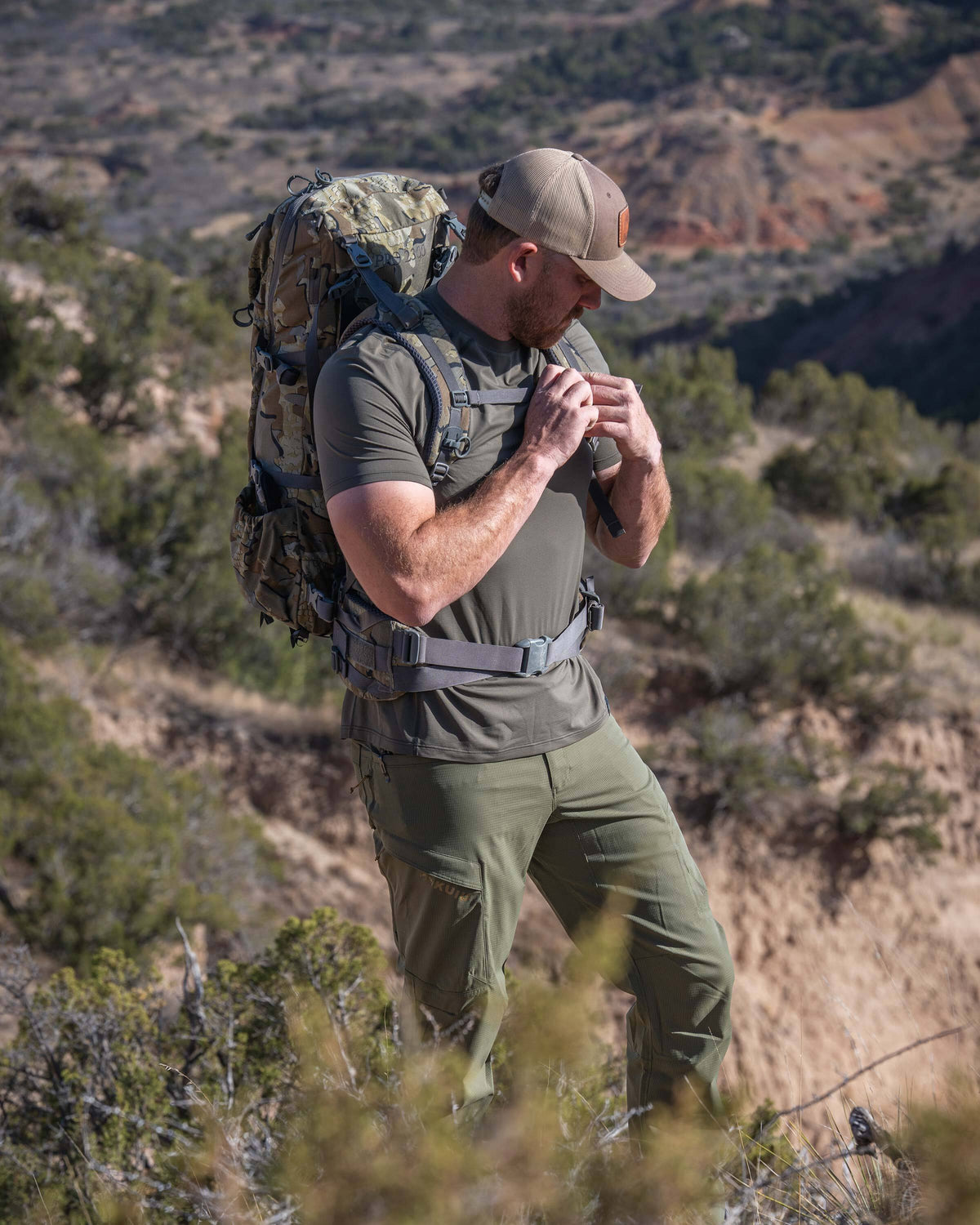 Hunter wearing Vital S1 Short Sleeve in Ash Brown, standing in an open rocky landscape with low shrubs and vegetation,  and fastening the chest straps on his pack. 