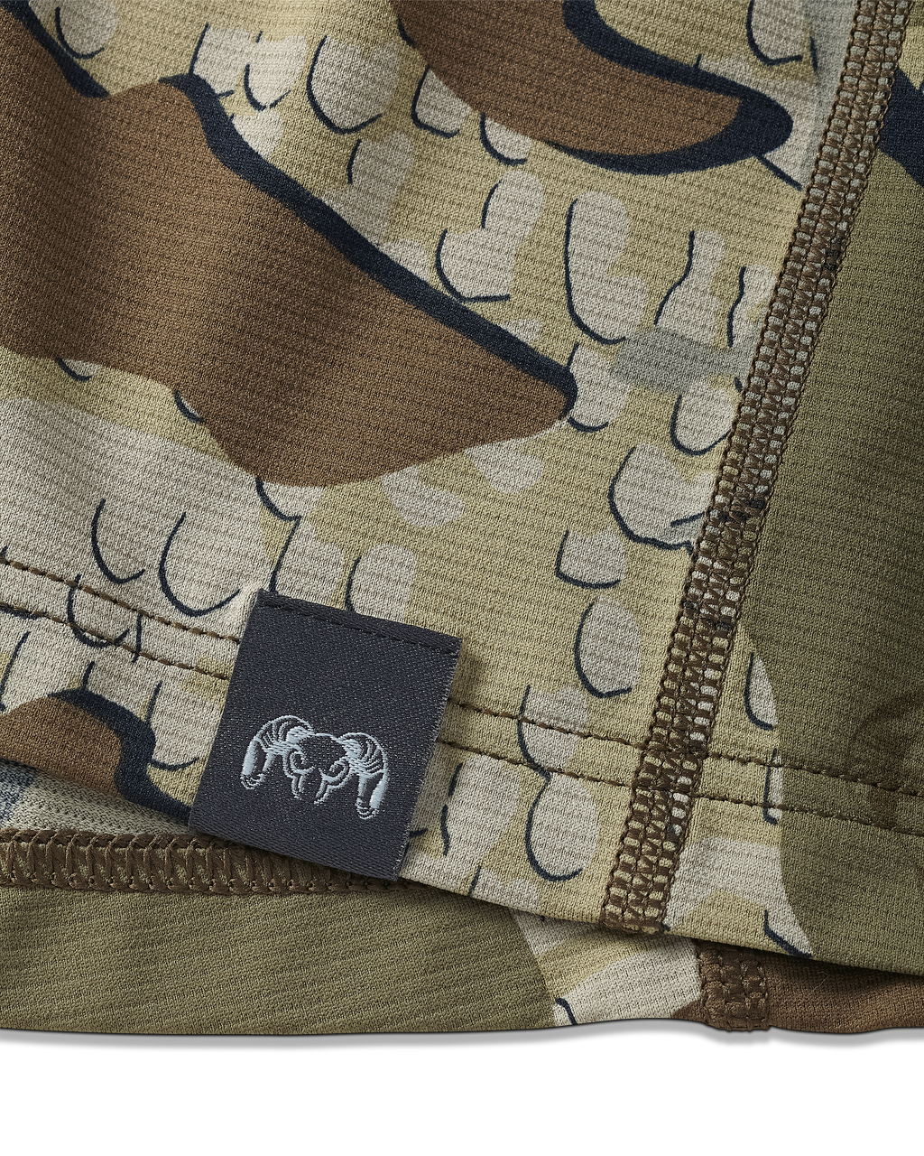 Close-up view of bottom seam of Vital S1 Short Sleeve in Valo Camouflage, Displaying a sewn in KUIU Ram logo on sewn-in tag 