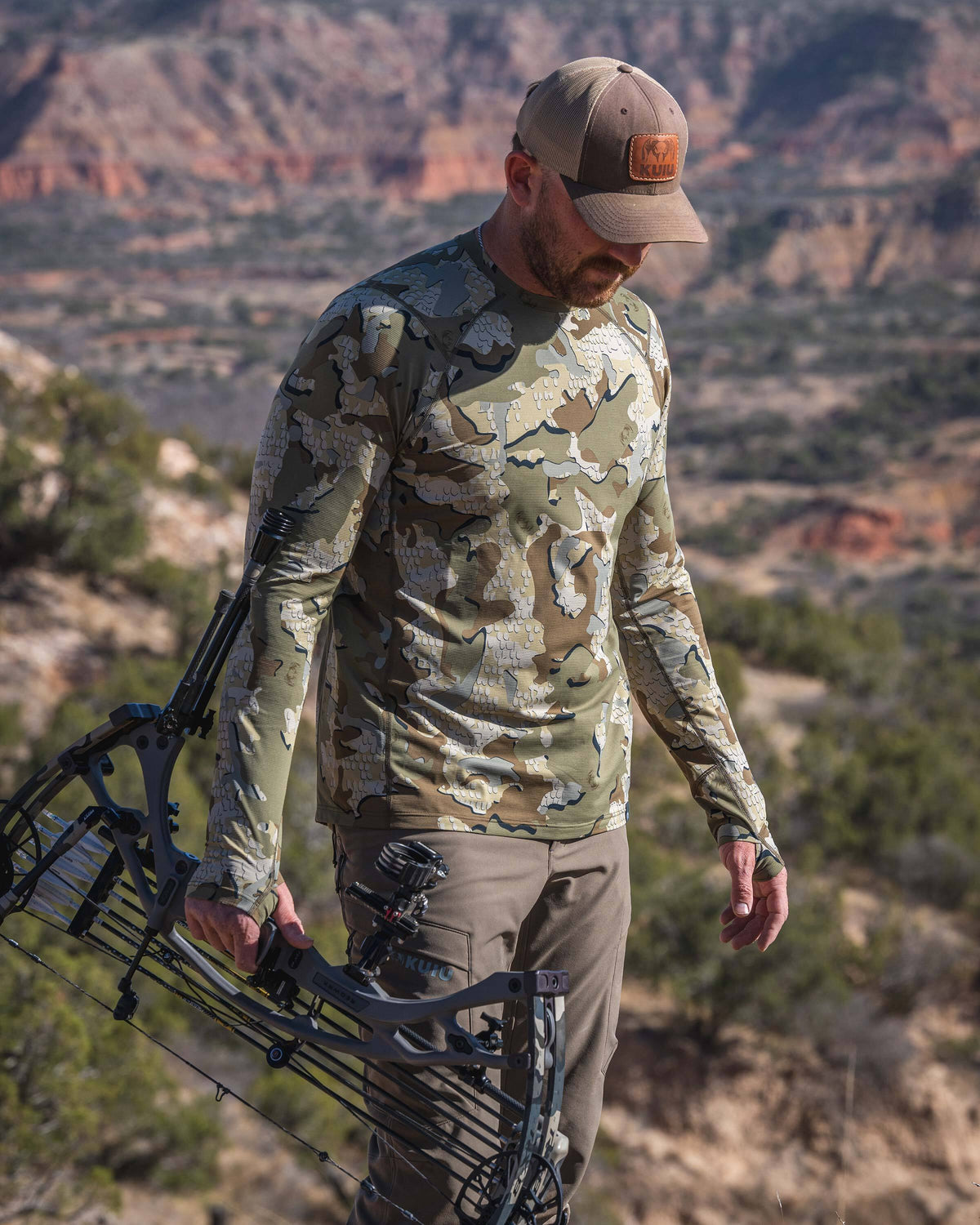 Hunter wearing Vital S1 Long Sleeve Crew in Valo Camouflage, holding a compound bow and standing on a rugged, rocky hillside.