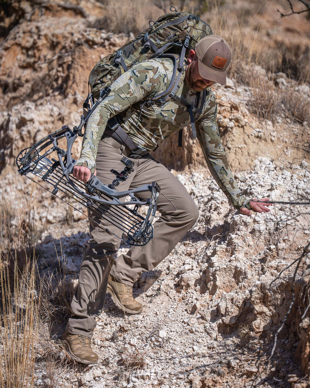 Hunter carefully stepping down a rocky, uneven slope in a dry, rugged outdoor landscape, wearing Vital S1 Long Sleeve Crew in Valo Camouflage.
