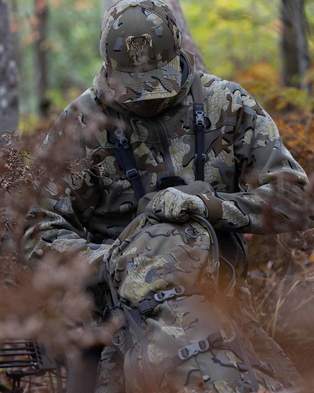 Hunter in the field wearing a Valo Camouflage Hat and the Peloton 240 Full Zip Hoodie in Valo Camouflage reaching into a backpack