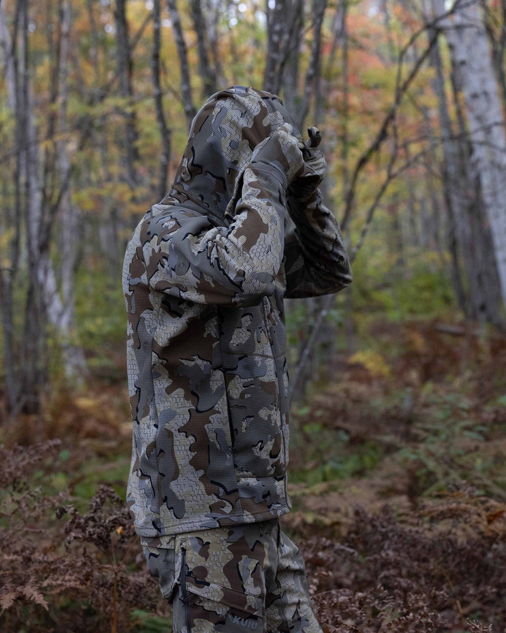 Hunter in the field wearing Valo Camouflage pants and the Peloton 240 Full Zip Hoodie in Valo Camouflage adjusting the hood