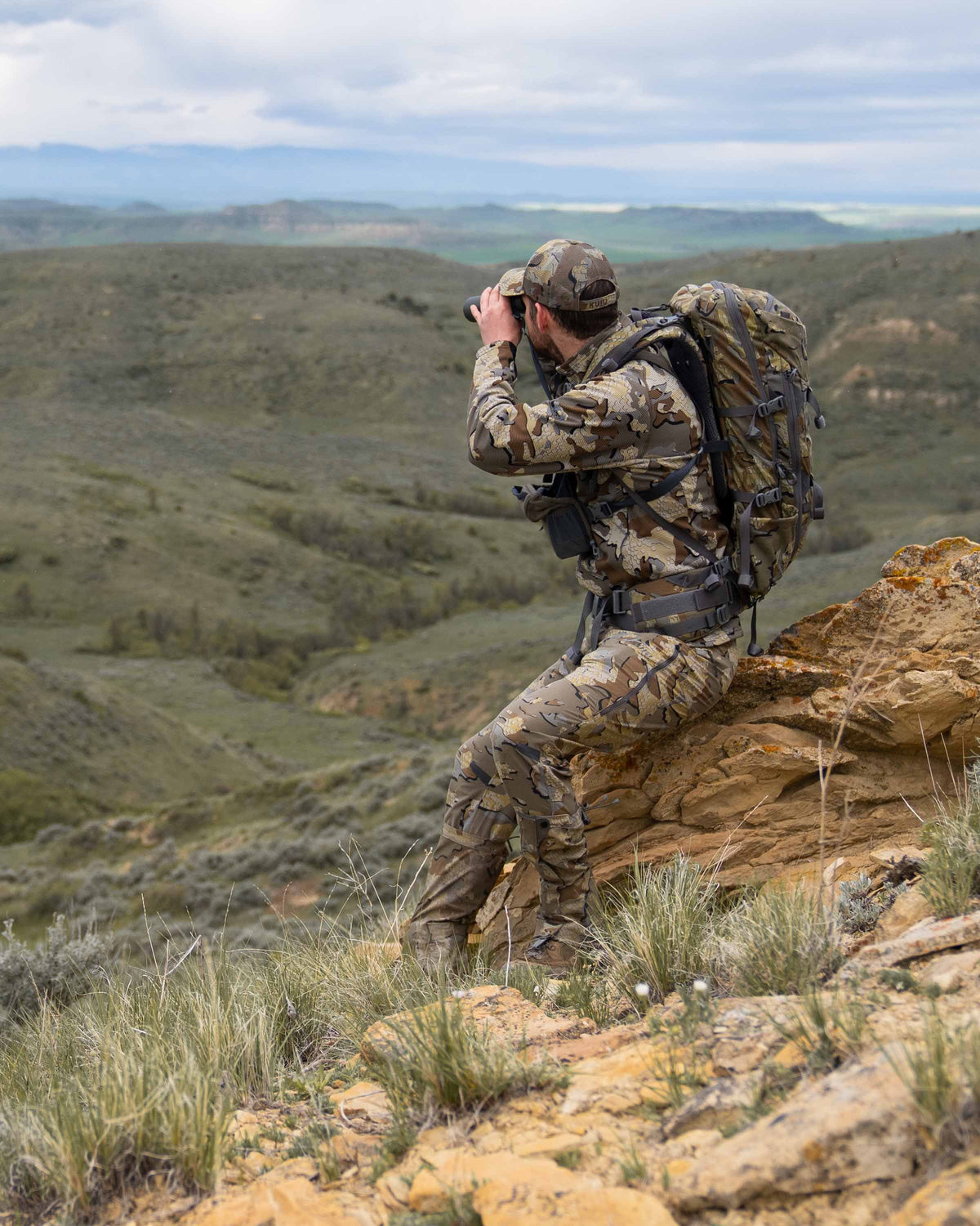 Hunter sitting on a rock wearing a full Valo kit including the Peloton 240 Full Zip in Valo Camouflage using binoculars
