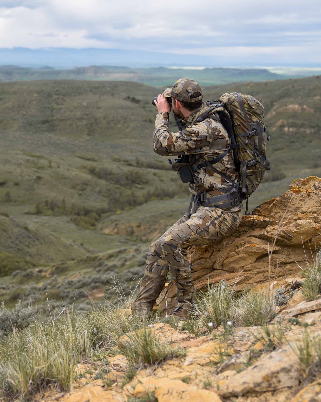 Hunter sitting on a rock wearing a full Valo kit including the Peloton 240 Full Zip in Valo Camouflage using binoculars