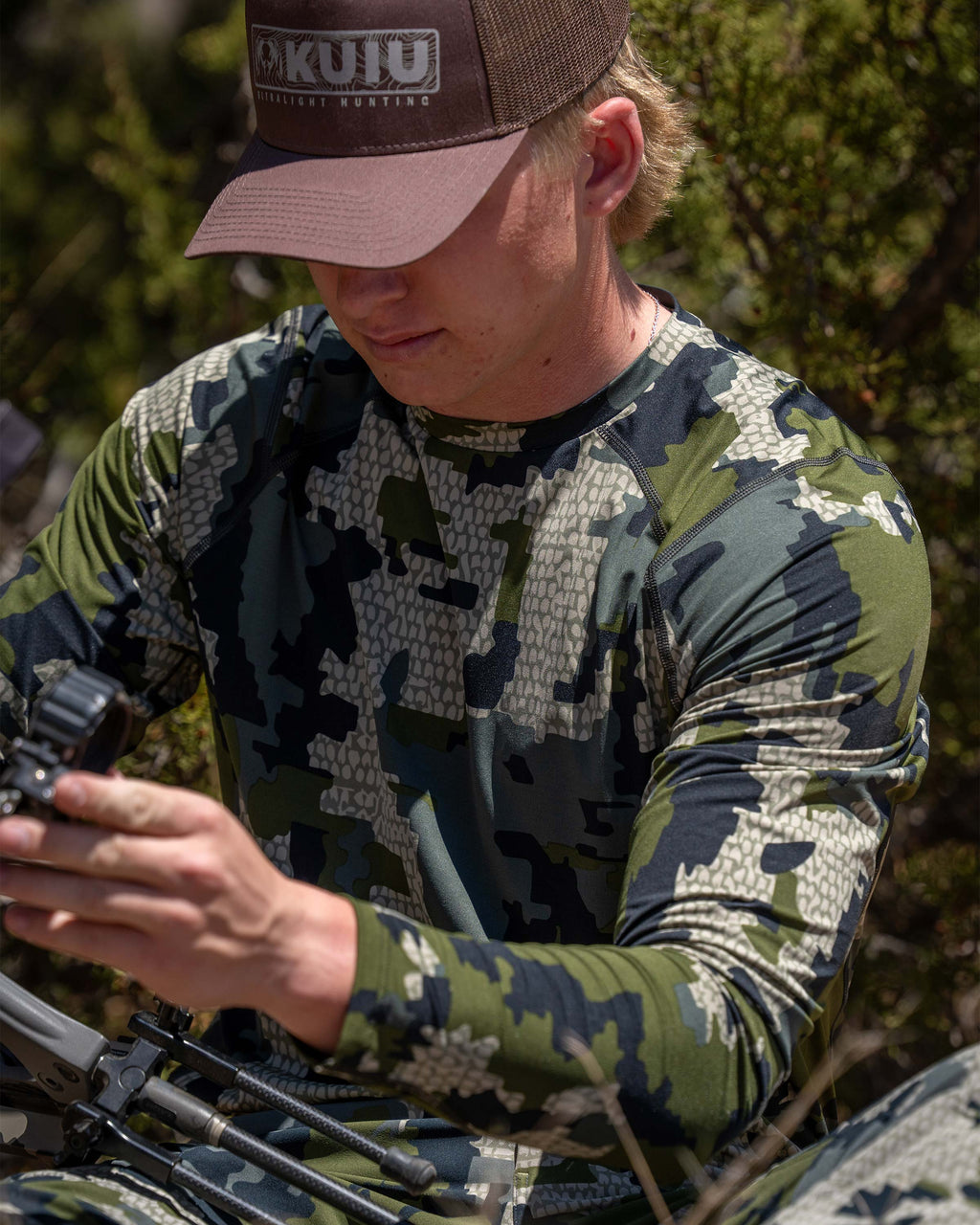 Hunter wearing Vital S2 Long Sleeve in Verde Camouflage adjusting the compound bow on his lap.