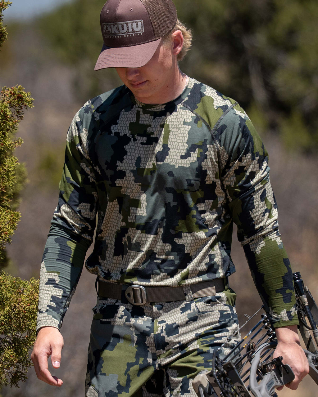 Hunter wearing Vital S2 Long Sleeve in Verde Camouflage holding a compound bow in a forest setting.