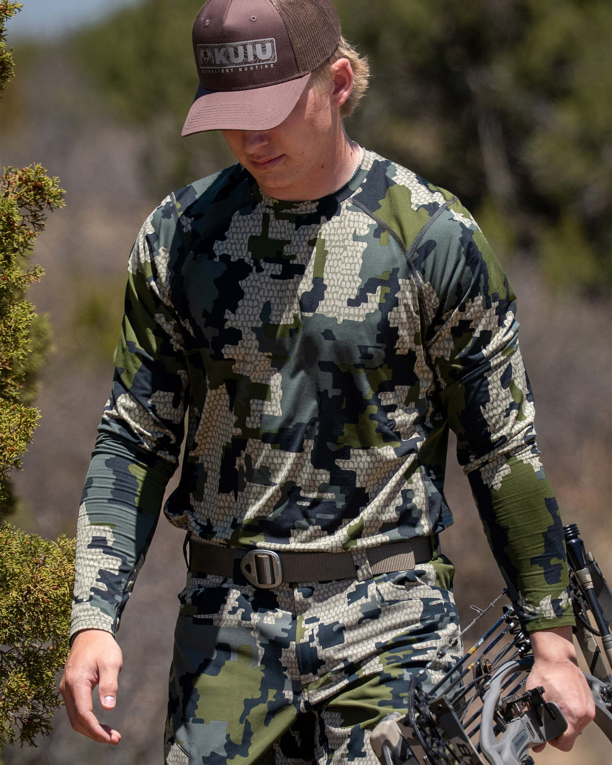 Hunter wearing Vital S2 Long Sleeve in Verde Camouflage holding a compound bow in a forest setting.