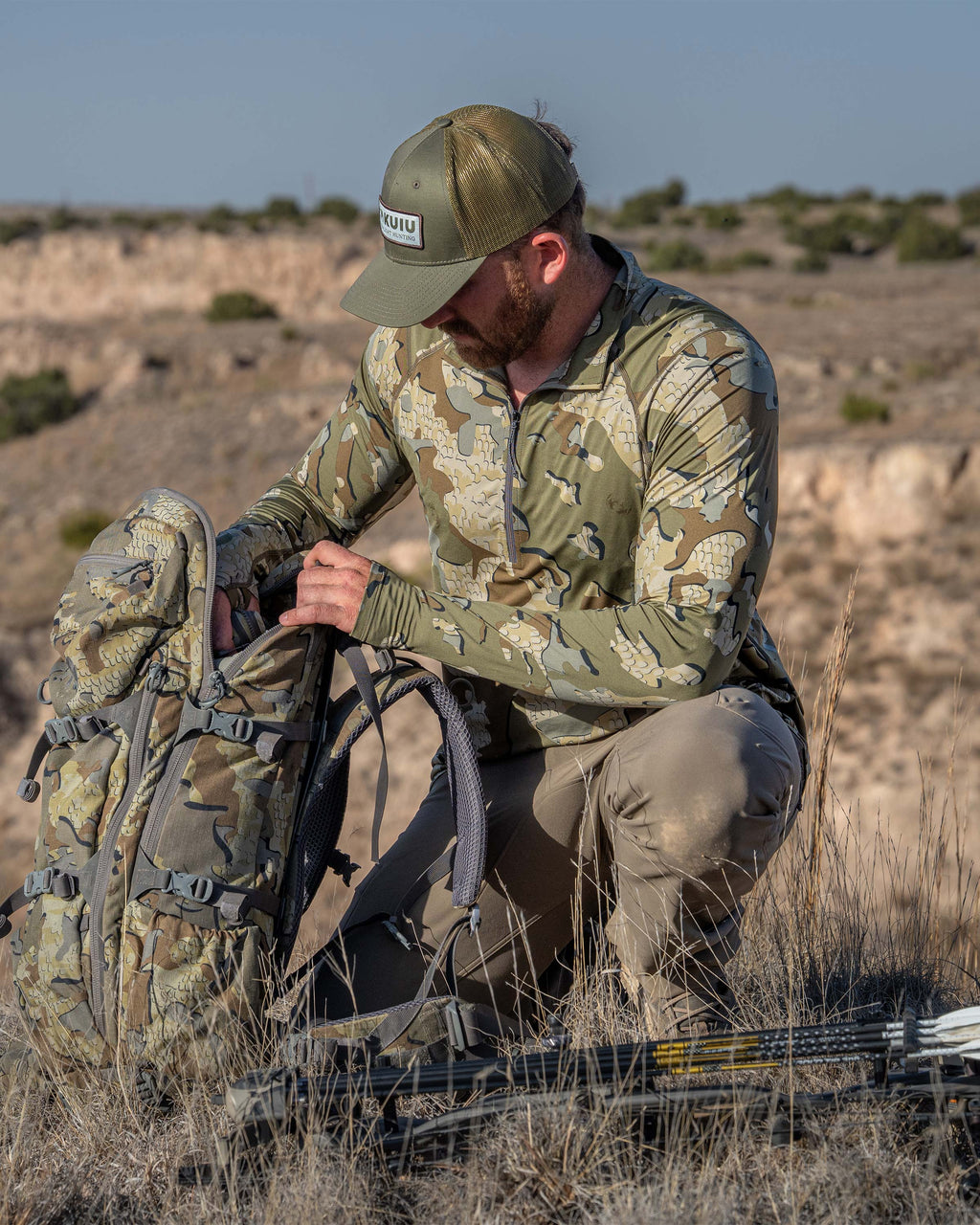 Hunter wearing Vital S2 Zip-T in Valo Camouflage kneeling in a desert landscape with a backpack and bow.