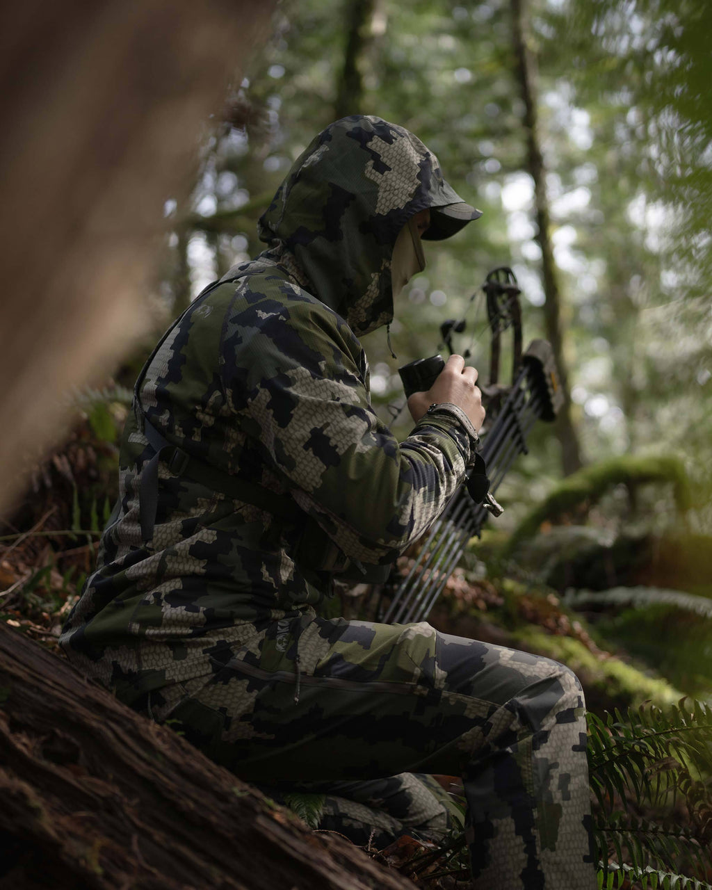 Hunter sitting down in the woods with his binocular and bow, wearing Tiburon Hybrid Hoodie in Verde Camouflage