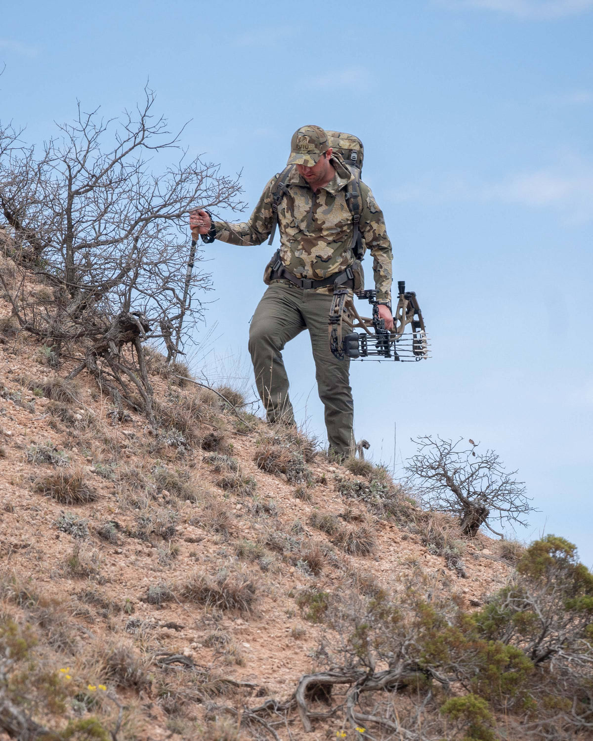 Hunter hiking uphill through a desert terrain landscape, wearing Tiburon Hybrid Hoodie in Valo Camouflage